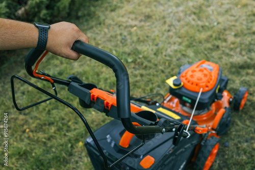 Close-up of a hand holding the handle of an orange lawn mower, seen from above. The mower is operating on a lawn, highlighting the details of the equipment and the action of mowing