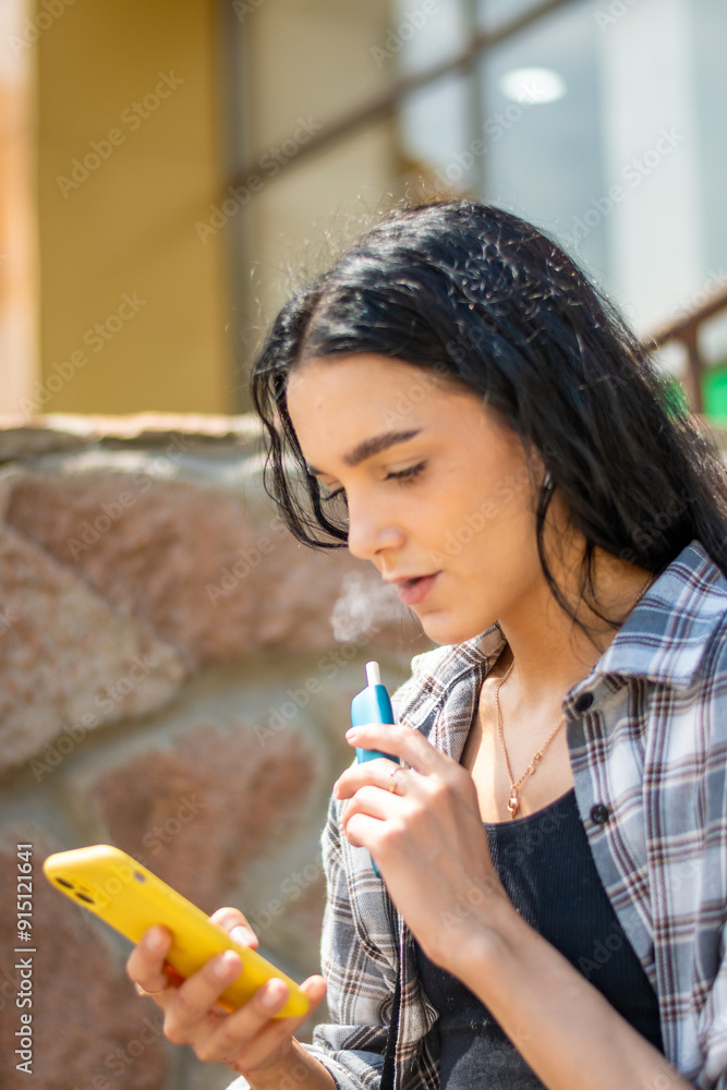Young woman smoking e-cigarette looking at the phone in the street. Addiction concept. High quality photo. Young woman smoking e-cigarette looking at the phone in the street. Addiction concept. High quality photo.