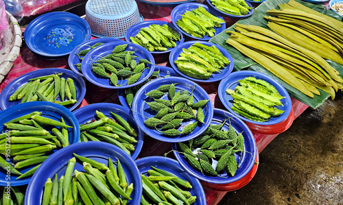 Assorted fresh green vegetables, including okra, bitter melon, and winged beans, displayed on blue plates at a market stall