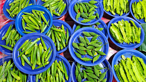 Top view of fresh okra, bitter melon, and winged beans neatly arranged on blue plates at a local market stall