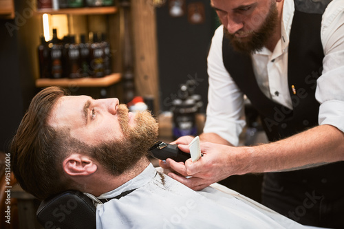 Professional barber using electric razor to shave beard. Regular client at an appointment in barbershop. Confident male customer getting his beard done.