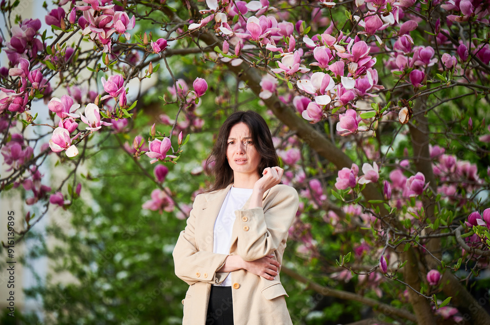 Woman allergic suffering from seasonal allergy at spring in blossoming garden at springtime. Young woman sneezing, having runny nose in front of blooming tree. Spring allergy concept Woman allergic suffering from seasonal allergy at spring in blossoming garden at springtime. Young woman sneezing, having runny nose in front of blooming tree. Spring allergy concept