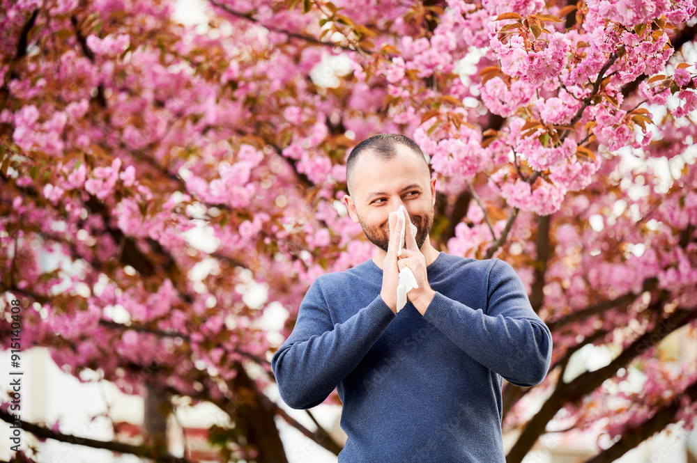 Man allergic suffering from seasonal allergy at spring in blossoming garden at springtime. Man sneezing and blowing nose using nasal handkerchief in front of blooming tree. Spring allergy concept. Man allergic suffering from seasonal allergy at spring in blossoming garden at springtime. Man sneezing and blowing nose using nasal handkerchief in front of blooming tree. Spring allergy concept.