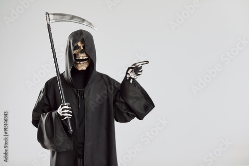 Konstfotografi Male person in Halloween grim reaper death costume holding scythe in black cloak with hood pointing index finger to the side isolated on studio gray background