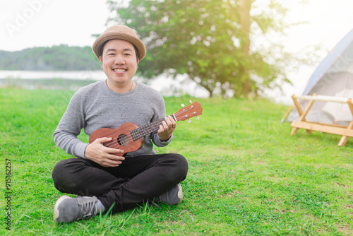 Cheerful Asian man playing ukulele while camping. Smiling musician in hat sitting on grass near tent, enjoying outdoor leisure activity. he relax and feeling happy, he training to artist.