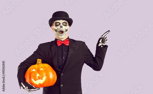 Shocked man dressed in Day of the Dead costume points his index finger to side on light lilac background. Man in black suit with skeleton painted on his face holds pumpkin and points to copy space. photo