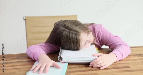 Junior schoolgirl clearly exhausted rests head on notebook. Small child with loose hair struggles to complete homework with tired eyes barely open