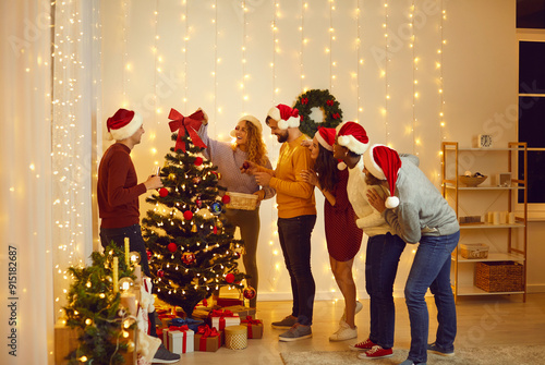Diverse group of happy people in Santa hats having a good time at Christmas party at home. Young mixed race friends decorating their tree with beautiful, stylish baubles in a cozy, modern living room
