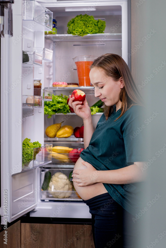 Pregnant woman is eating an apple near the refrigerator Pregnant woman is eating an apple near the refrigerator