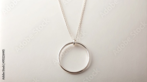 A delicate, silver thin circle pendant hangs from a dainty chain, suspended against a soft, creamy white background, evoking a sense of simplicity and elegance.