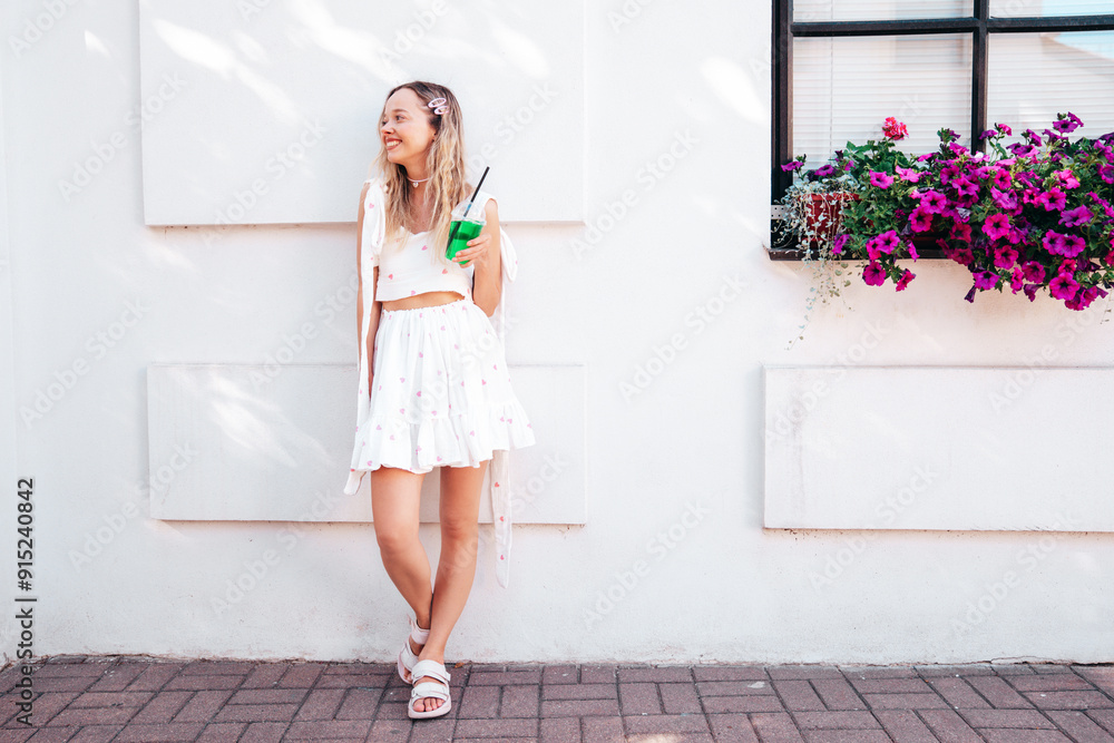 Young beautiful smiling hipster woman in trendy summer clothes. Sexy carefree model posing in the street. Holding and drinking fresh vegetable cocktail drink in plastic cup with straw. In sunny day Young beautiful smiling hipster woman in trendy summer clothes. Sexy carefree model posing in the street. Holding and drinking fresh vegetable cocktail drink in plastic cup with straw. In sunny day