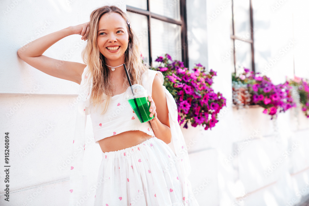 Young beautiful smiling hipster woman in trendy summer clothes. Sexy carefree model posing in the street. Holding and drinking fresh vegetable cocktail drink in plastic cup with straw. In sunny day Young beautiful smiling hipster woman in trendy summer clothes. Sexy carefree model posing in the street. Holding and drinking fresh vegetable cocktail drink in plastic cup with straw. In sunny day