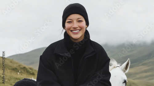 A woman smiles while wearing a black beanie and coat, sitting outdoors on a white horse with a mountainous background on a cloudy day. photo