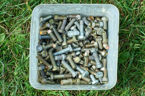 many iron new small threaded shiny identical industrial bolts in plastic white box stands on green grass during daytime outside photo