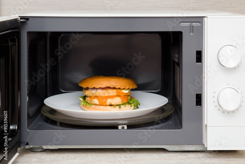 Fresh big burger on white plate in opened microwave oven at home kitchen. Food warm up. Closeup. Front view.