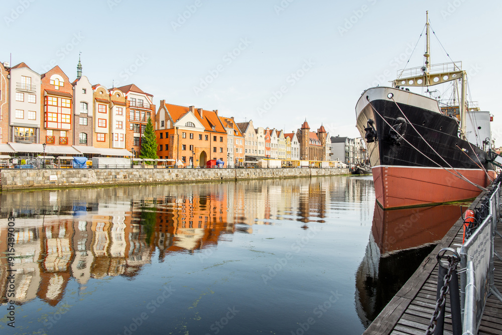 Fototapeta premium A historic black and red ship is docked along the Motlawa River in Gdansk, Poland, with picturesque buildings reflected in the water.