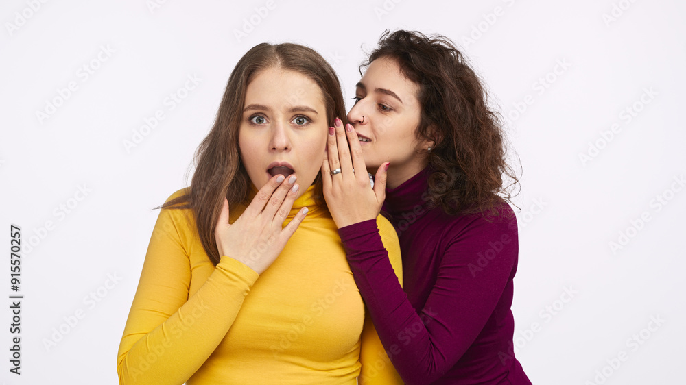 Two women, one in a yellow shirt and the other in a purple shirt, sharing a secret. The woman in yellow shows a surprised expression. Two women, one in a yellow shirt and the other in a purple shirt, sharing a secret. The woman in yellow shows a surprised expression.