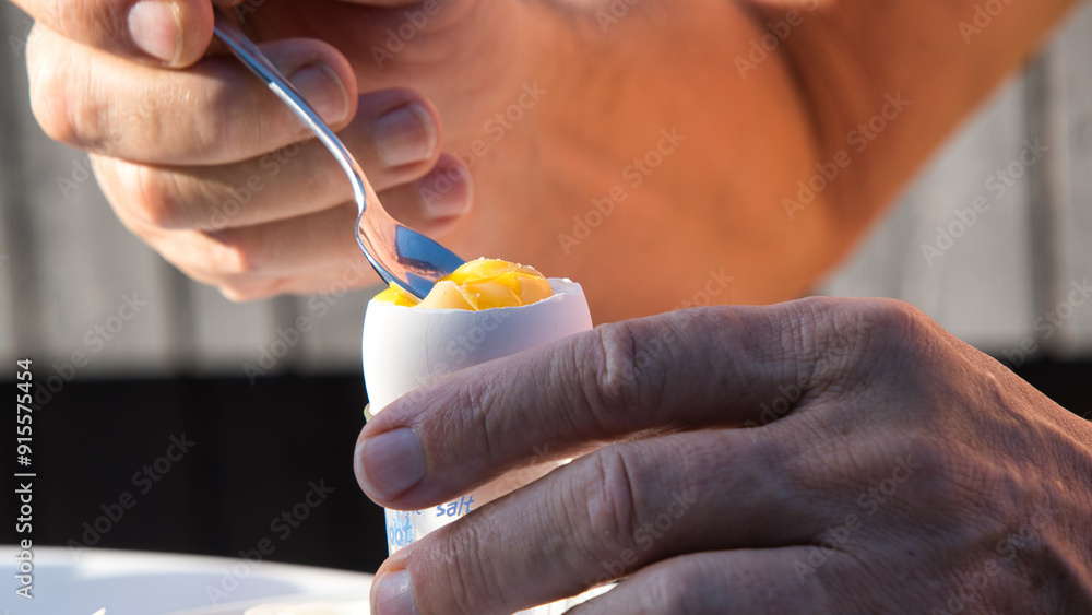 A person is scooping a yellow egg yolk from soft boiled egg with shell during breakfast time. A person is scooping a yellow egg yolk from soft boiled egg with shell during breakfast time.