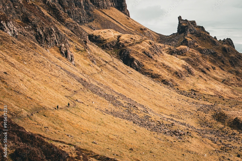 Fototapeta premium Hikers walking on a trail in a mountainous landscape with rugged terrain and a cloudy sky.