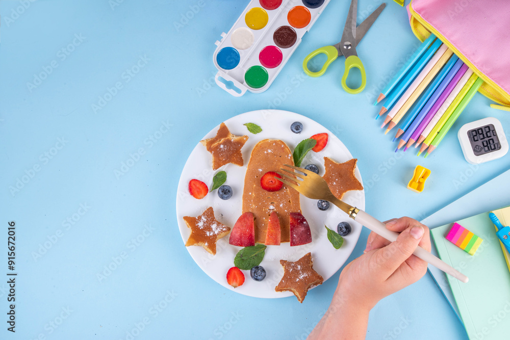 Naklejka premium Back to school, school kids breakfast with creative shaped pancakes and fruit berries, on light blue background with school supplies, top view copy space. Kids hands eating pancakes