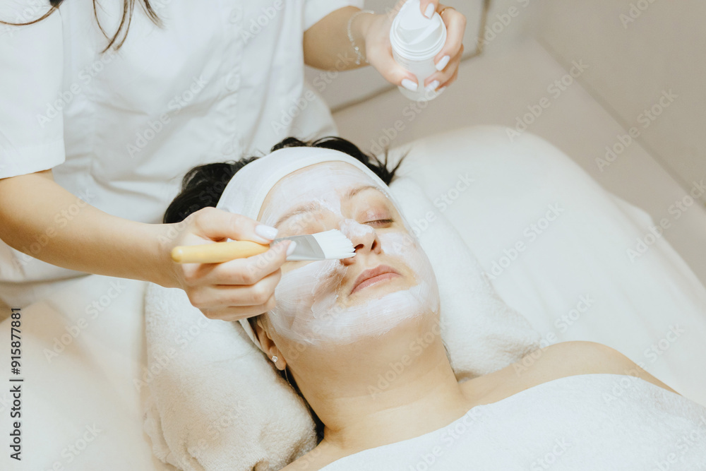 A girl cosmetologist applies a cream mask with a brush to a woman s face. A girl cosmetologist applies a cream mask with a brush to a woman s face.