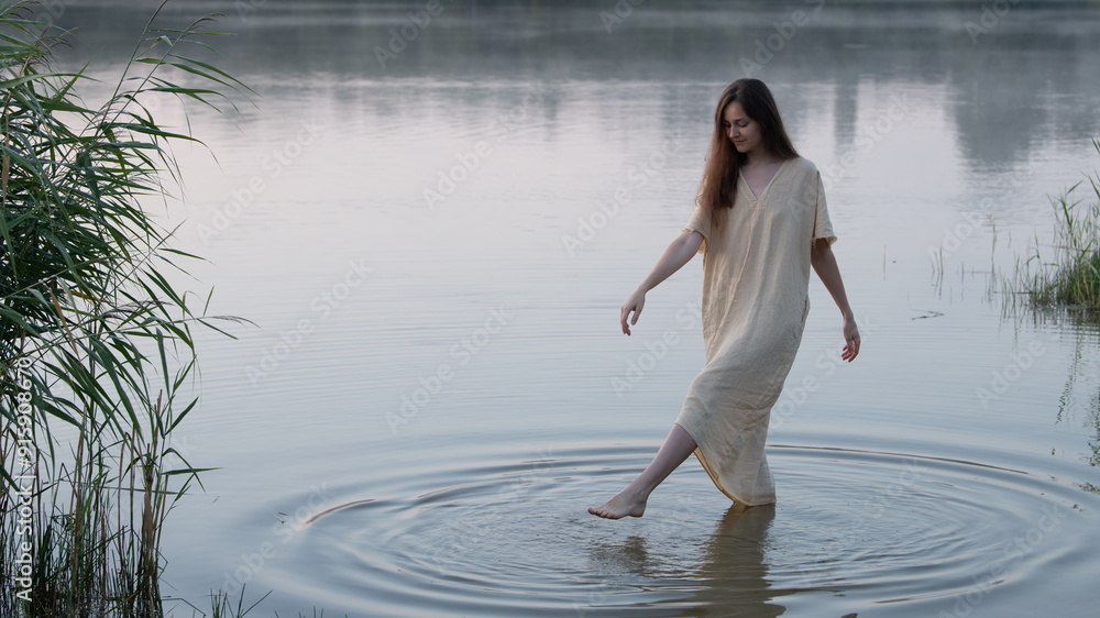 Fototapeta premium A dreamy girl or young woman in a simple light dress draws circles in the water with her feet in the middle of a lake or river