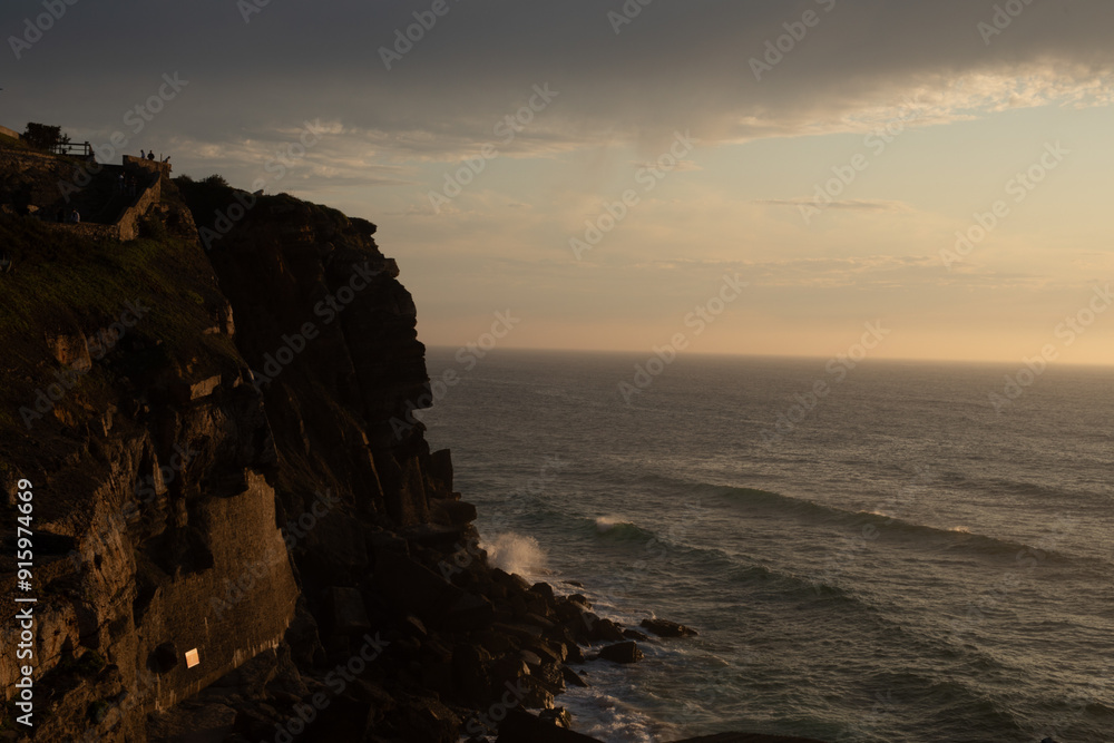 A dramatic cliff face stands tall against a soft sunset sky overlooking the vast ocean below A dramatic cliff face stands tall against a soft sunset sky overlooking the vast ocean below