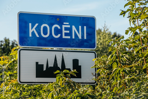 A town sign for Kocēni, featuring a blue and white design with the town name in large letters. Below the name is a silhouette of a cityscape, surrounded by lush green foliage. The sign is set against photo