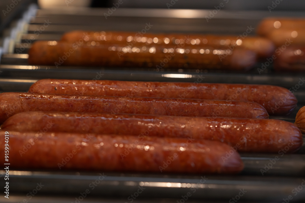 A close-up of several hot dogs being cooked on a rotating roller grill. The sausages glisten with a savory sheen, highlighting their perfectly grilled texture. The background shows a busy kitchen A close-up of several hot dogs being cooked on a rotating roller grill. The sausages glisten with a savory sheen, highlighting their perfectly grilled texture. The background shows a busy kitchen