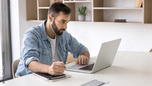 Focused handsome Latin business professional man in casual working at laptop in office, writing notes, sitting at workplace table, watching online video, learning seminar, using technology