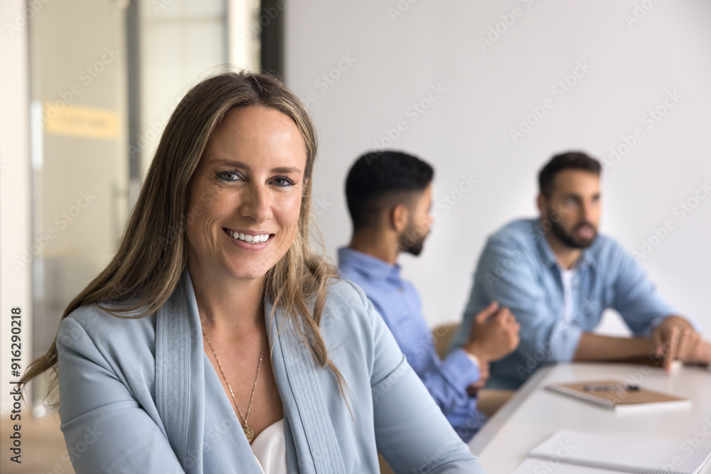 Happy positive confident business leader woman sitting at meeting table, posing for professional portrait with diverse colleagues talking in background, looking at camera, smiling Happy positive confident business leader woman sitting at meeting table, posing for professional portrait with diverse colleagues talking in background, looking at camera, smiling