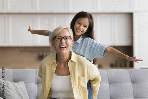 Cuadro en lienzo Active blonde grandma holding sweet beautiful kid girl from behind, looking at camera with toothy smile, laughing, enjoying being grandmother