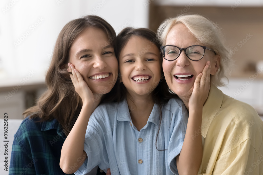 Happy kid girl touching cheeks of young mom and senior grandma, looking at camera with toothy smile. Positive cute child, mother and grandmother sitting close, posing for family portrait Happy kid girl touching cheeks of young mom and senior grandma, looking at camera with toothy smile. Positive cute child, mother and grandmother sitting close, posing for family portrait