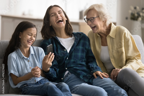 Excited girls and women of three family generations using mobile phone, laughing out loud at funny online content, enjoying online communication on home couch together