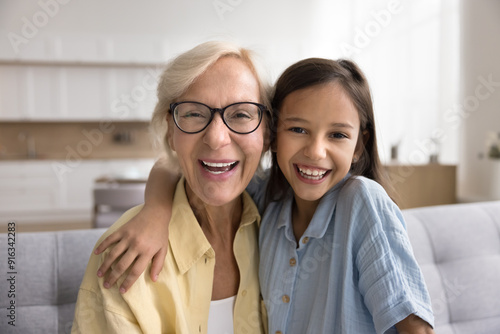 Positive grandmother in glasses and cute grandkid girl hugging, looking at camera, talking on video call at home, posing for family portrait with toothy smile, laughing. Grandma and kid head shot