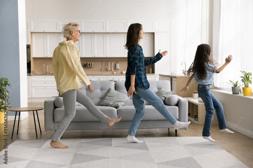 Εκτύπωση καμβά Cheerful girl, young mother and grandma dancing together in living room, doing walking movements, having fun at home party, enjoying family activity, leisure