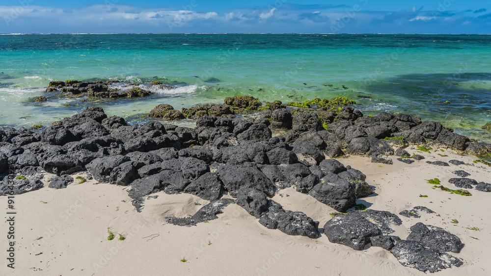 The picturesque seacoast. There is a pile of black volcanic lava boulders on the white sandy beach. Algae on the rocks. Turquoise ocean to the horizon. Blue sky, clouds. Mauritius. Belle Mare. The picturesque seacoast. There is a pile of black volcanic lava boulders on the white sandy beach. Algae on the rocks. Turquoise ocean to the horizon. Blue sky, clouds. Mauritius. Belle Mare.