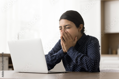 Young Indian woman feeling sleepy or fatigued, looks bored or disengaged with her current task, sits at table with laptop, cover mouth with palms while yawning, sign of natural drowsiness, tiredness photo