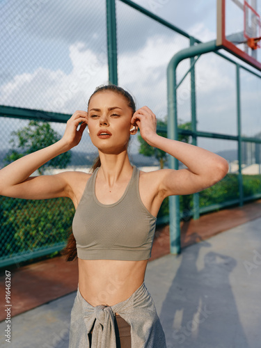 Young woman in sportswear adjusting earphones on an outdoor basketball court with green fencing and cloudy sky photo