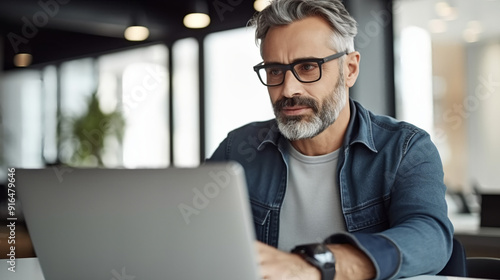 Portrait of an attractive mature man with glasses, grey hair and short beard sitting in front of his laptop working at the office.