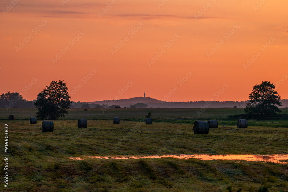 Beautiful rural landscape. A wet meadow with hay bales in the foreground. A hill with a lighthouse in the distance. The sky lit by the setting sun for a moment after the storm