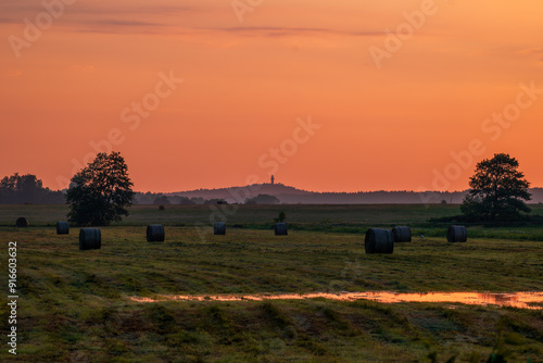 Obraz na płótnie Beautiful rural landscape. A wet meadow with hay bales in the foreground. A hill with a lighthouse in the distance. The sky lit by the setting sun for a moment after the storm w salonie