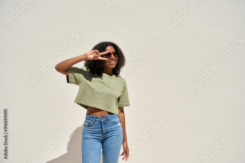 Happy joyful playful young African American woman wearing sunglasses, top and jeans having fun looking away standing at white wall background. Funky gen z Black girl celebrating sunny summer party.