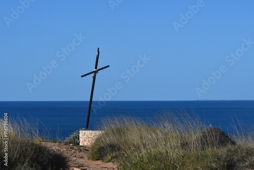 A wooden cross blackened with age and tied together with rope stands on a deserted headland. It rests on sandstone, and is surrounded by wild grasses. It is seen against a blue sea and sky.