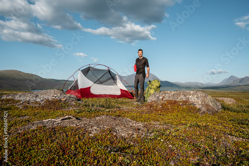 Smiling male white young man building a tent on a kiking adventure in swedish Padjelanta mountain National Park close to Kutjaurestugan holding a red bottle photo