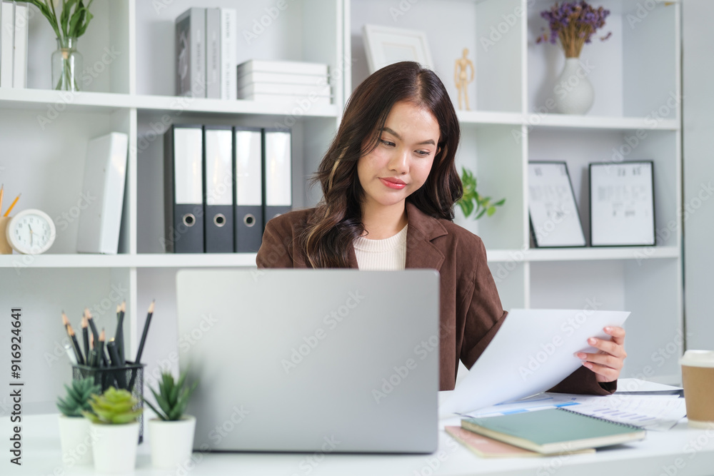 Young businesswoman in a brown blazer and beige sweater is seated at her desk, intently reviewing documents while working on her laptop in a modern home office. Young businesswoman in a brown blazer and beige sweater is seated at her desk, intently reviewing documents while working on her laptop in a modern home office.