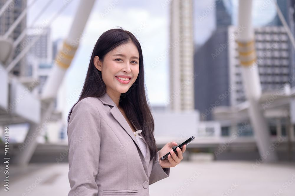 A confident young businesswoman stands outdoors in a modern urban setting, smiling while holding her smartphone. A confident young businesswoman stands outdoors in a modern urban setting, smiling while holding her smartphone.