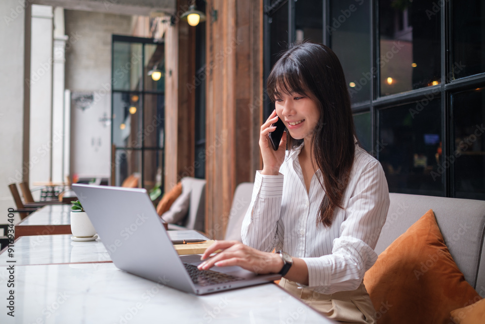 Business woman is seated at an outdoor café table, engaged in a phone conversation while working on her laptop. Business woman is seated at an outdoor café table, engaged in a phone conversation while working on her laptop.