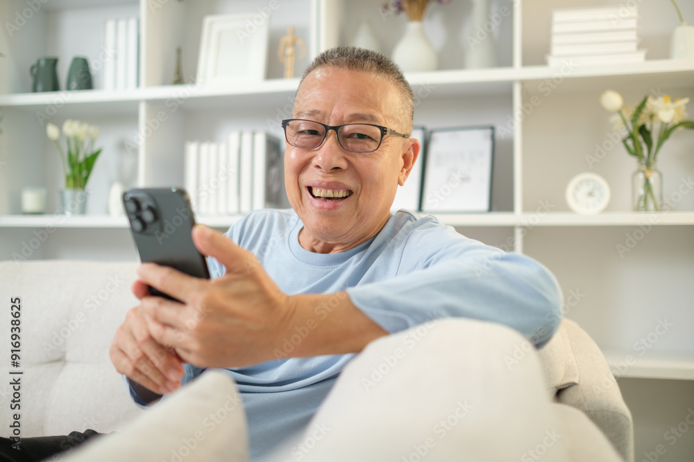 Smiling elderly man wearing glasses enjoying his time on the device, using mobile phone on sofa at home. Smiling elderly man wearing glasses enjoying his time on the device, using mobile phone on sofa at home.