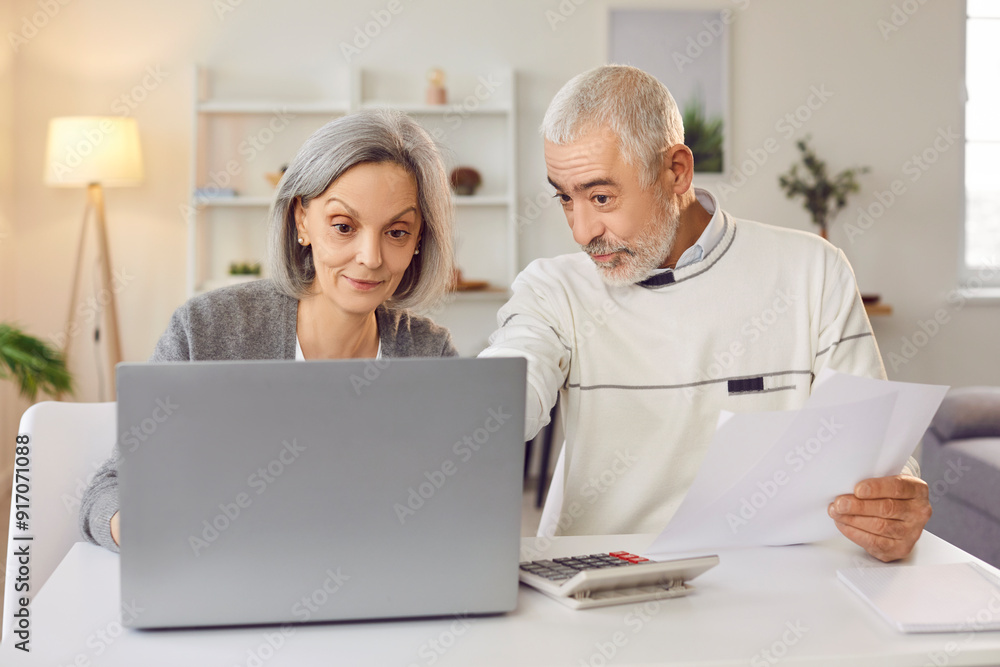Family documentation. Mature couple immersed in management of their home budget using modern technology. Mature husband and wife sitting at home at table with laptop, calculator and paper documents. Family documentation. Mature couple immersed in management of their home budget using modern technology. Mature husband and wife sitting at home at table with laptop, calculator and paper documents.